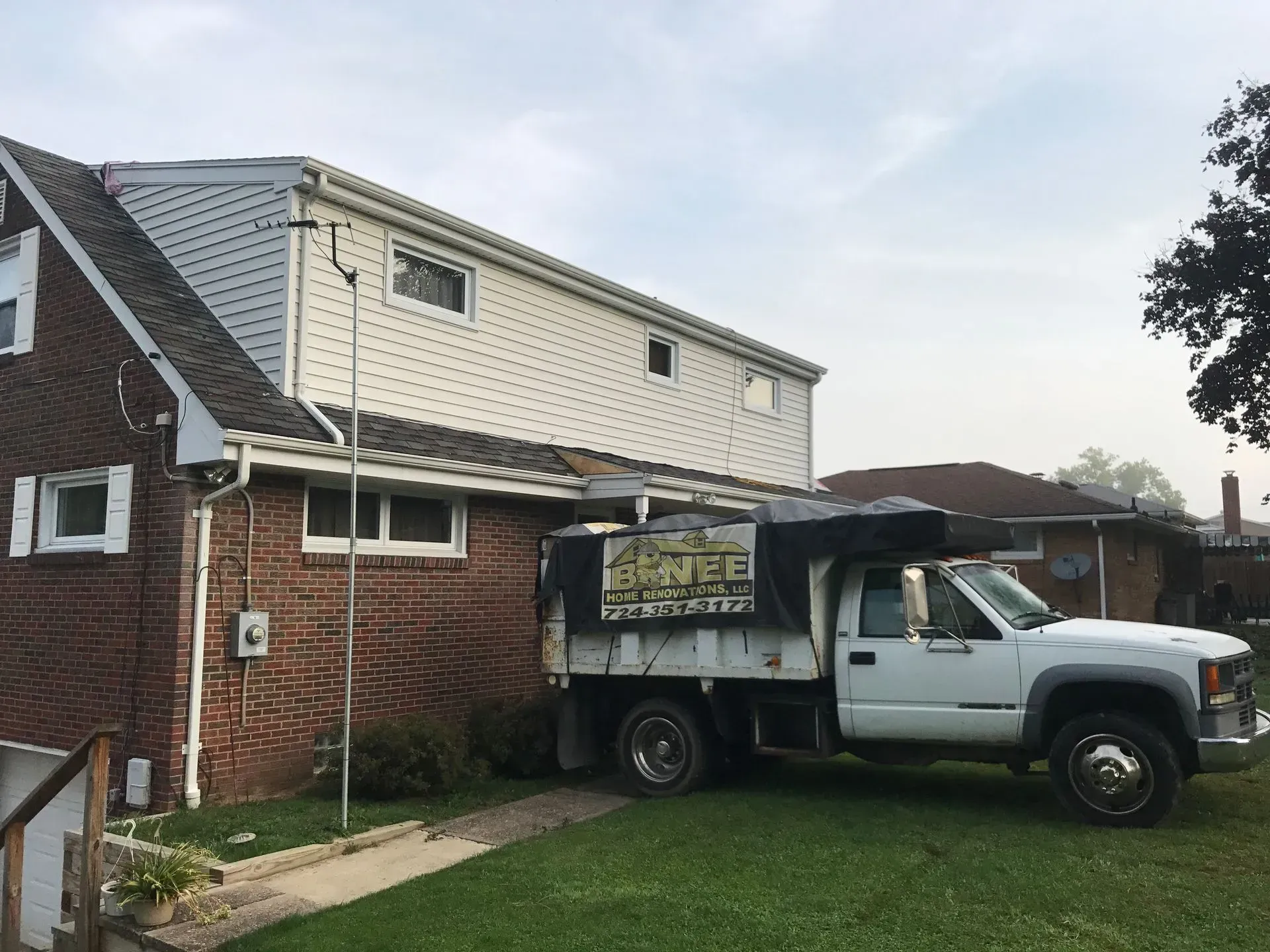 White truck parked next to a brick house with a white upper story, on a green lawn.