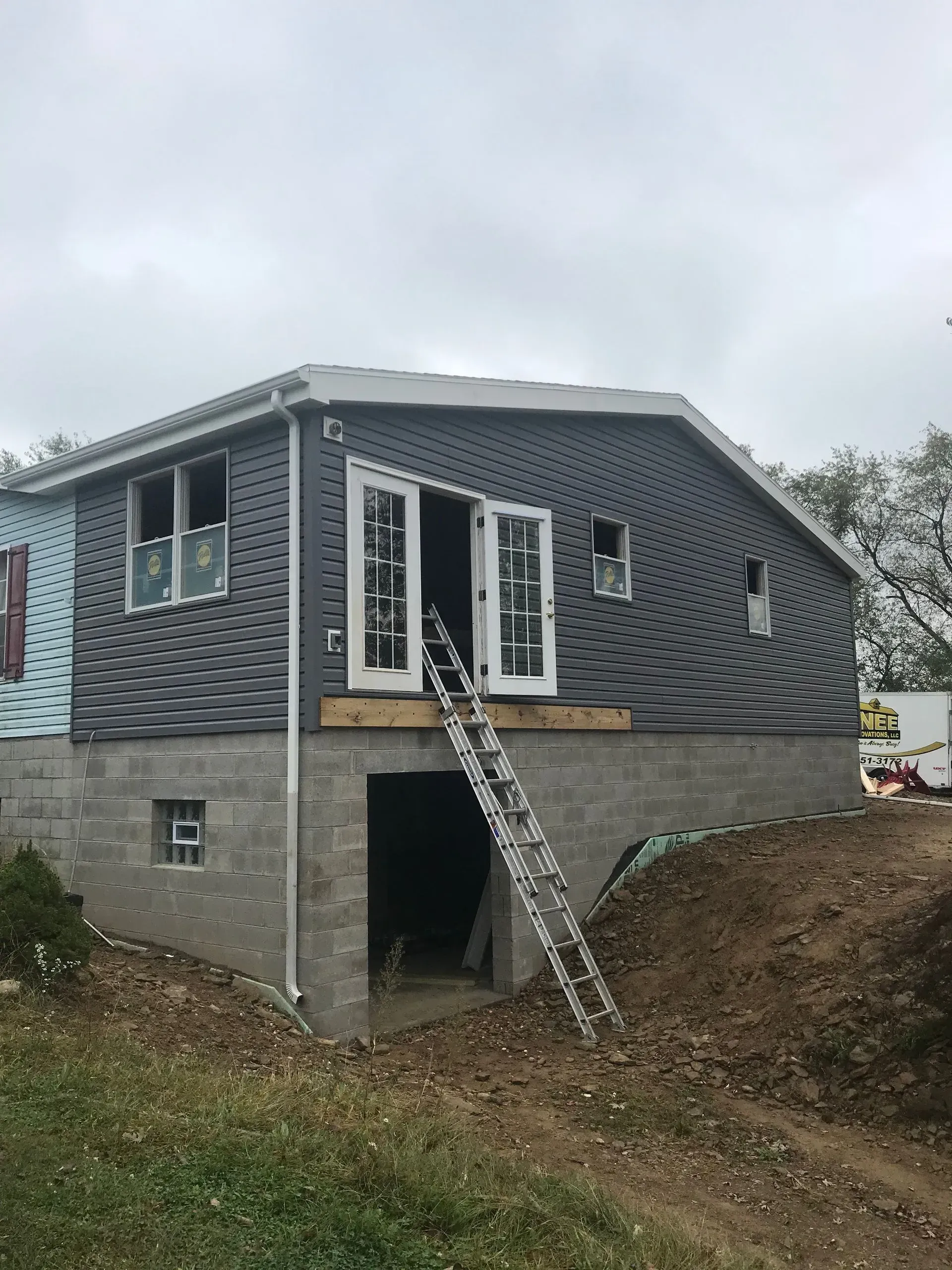 Two-story building under construction with gray siding and French doors. A ladder leans into the open doorway.