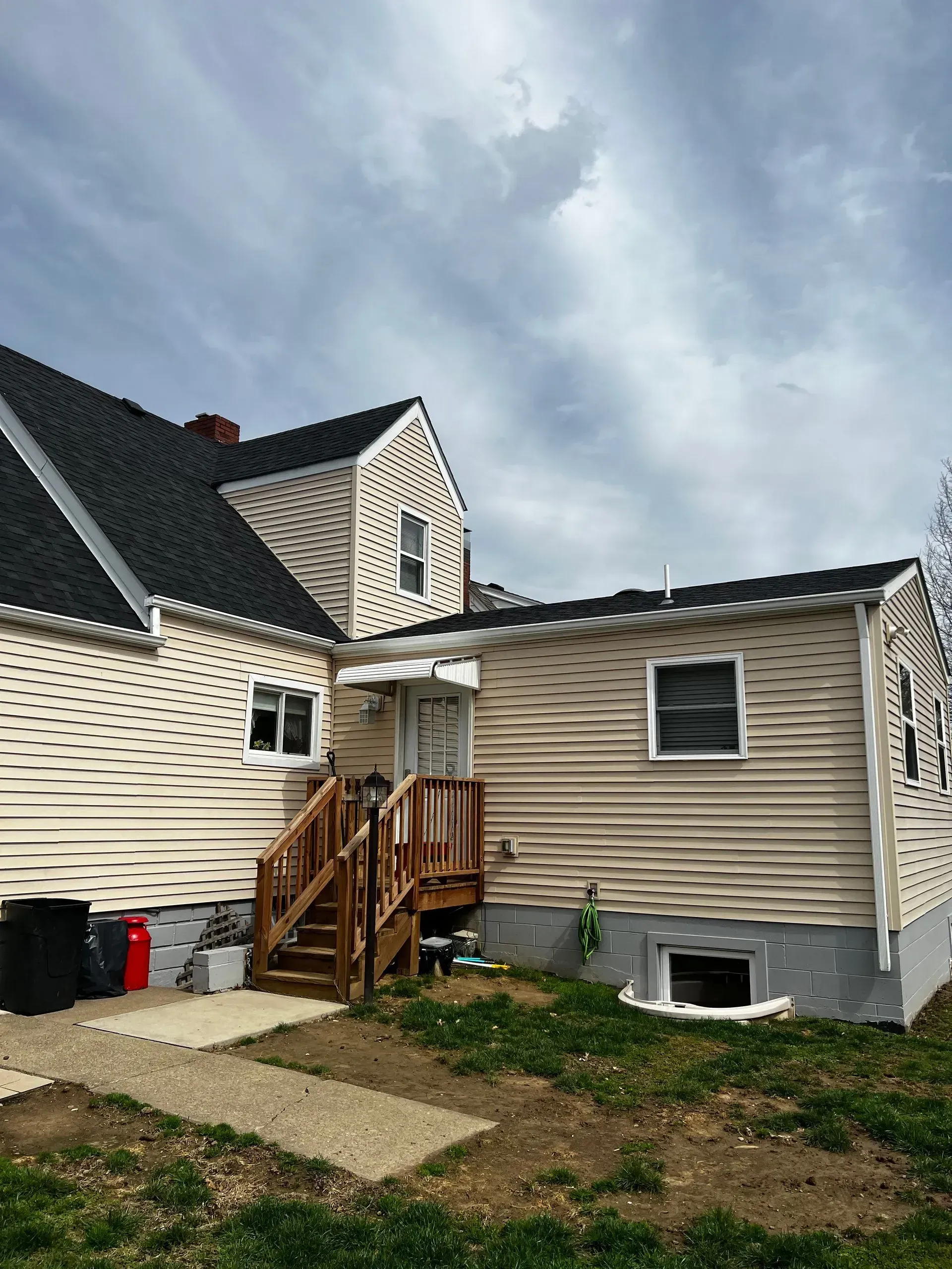 Back of a tan house with a dark roof and a wooden deck, under a cloudy sky.