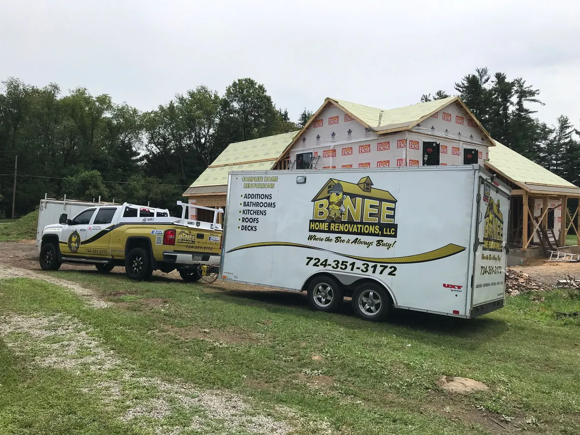 A yellow and white work truck towing a trailer in front of a house under construction.