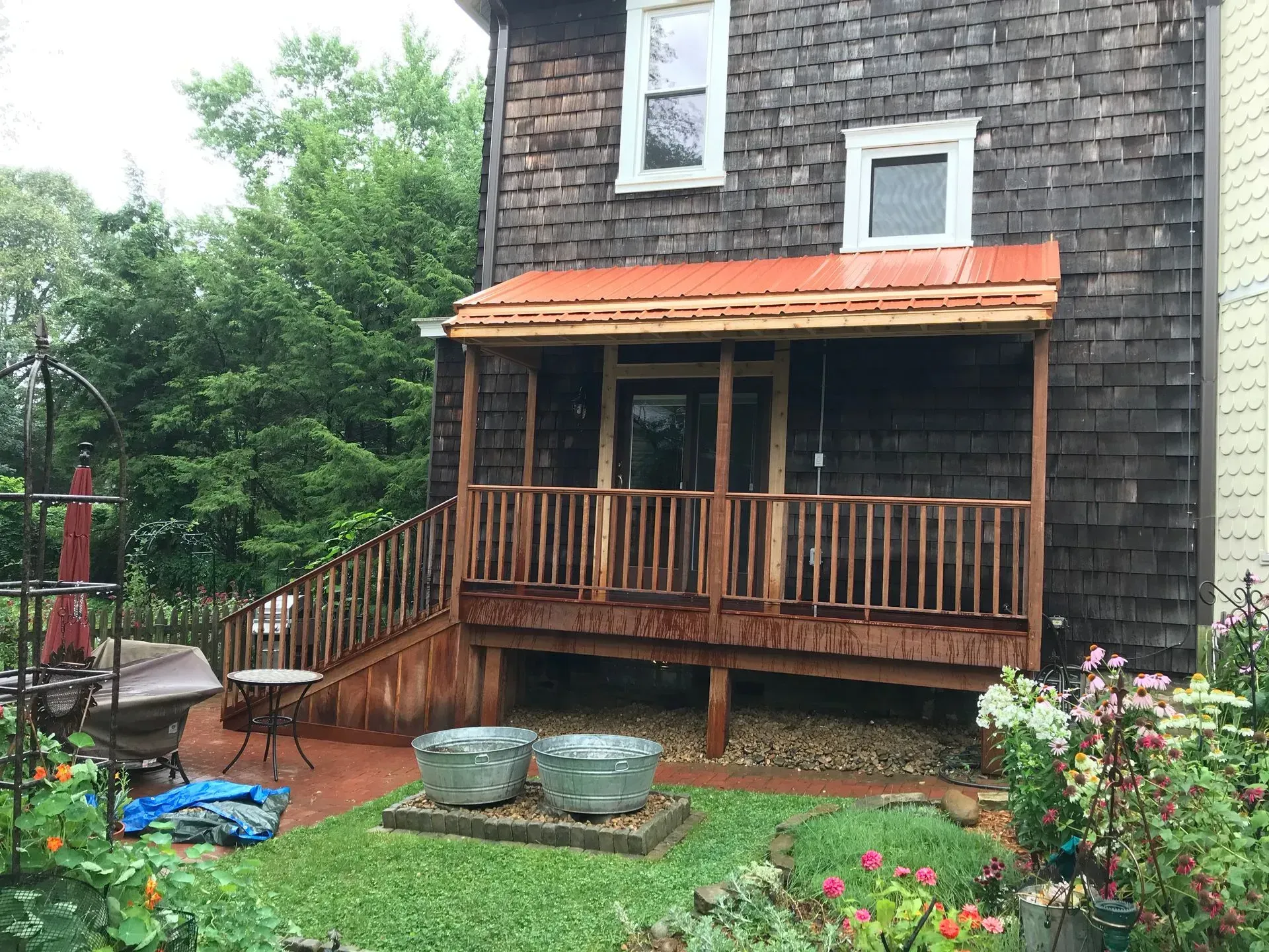 A wooden porch with a red roof on the side of a brown-shingled building, surrounded by a garden.