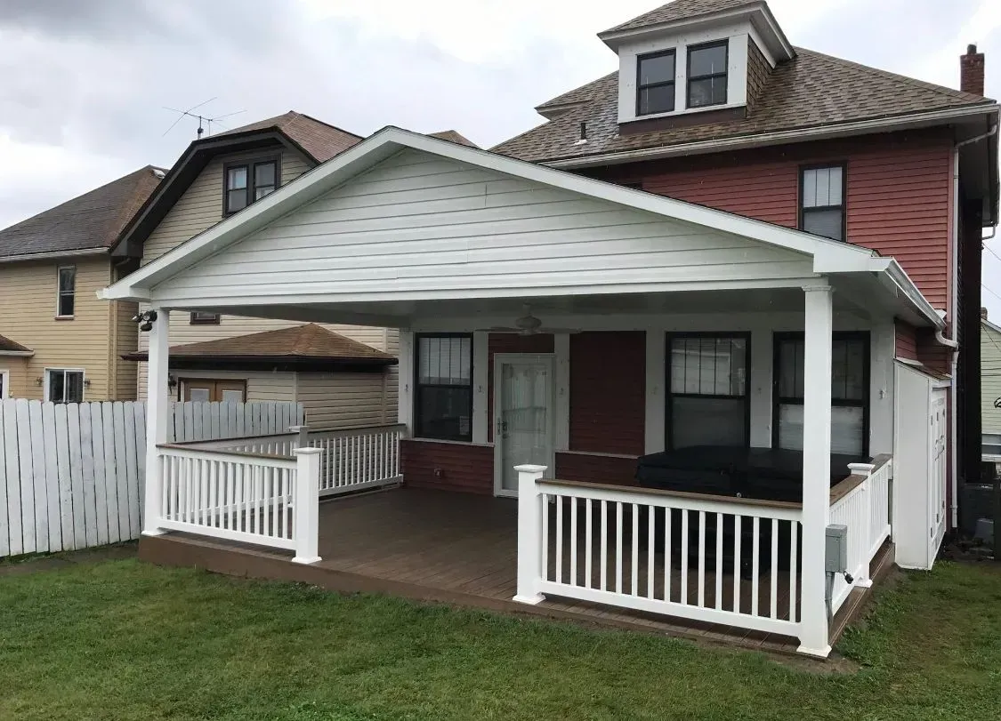 A two-story red brick house with a white-railed deck and a covered patio on a grassy lawn.