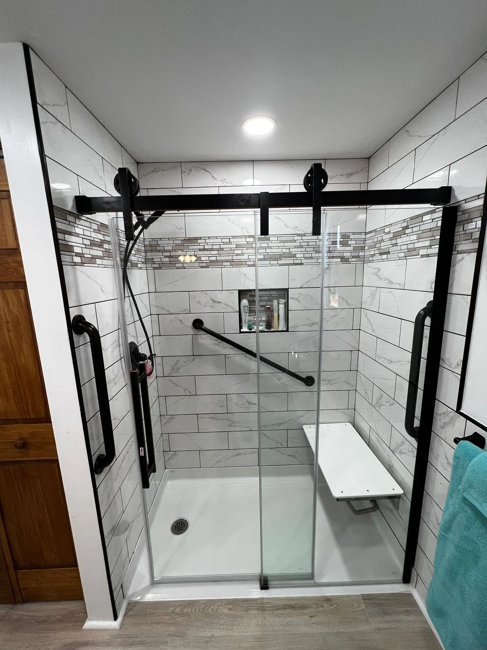 Modern walk-in shower with white tile, black trim, glass doors, a built-in bench, and grab bars.