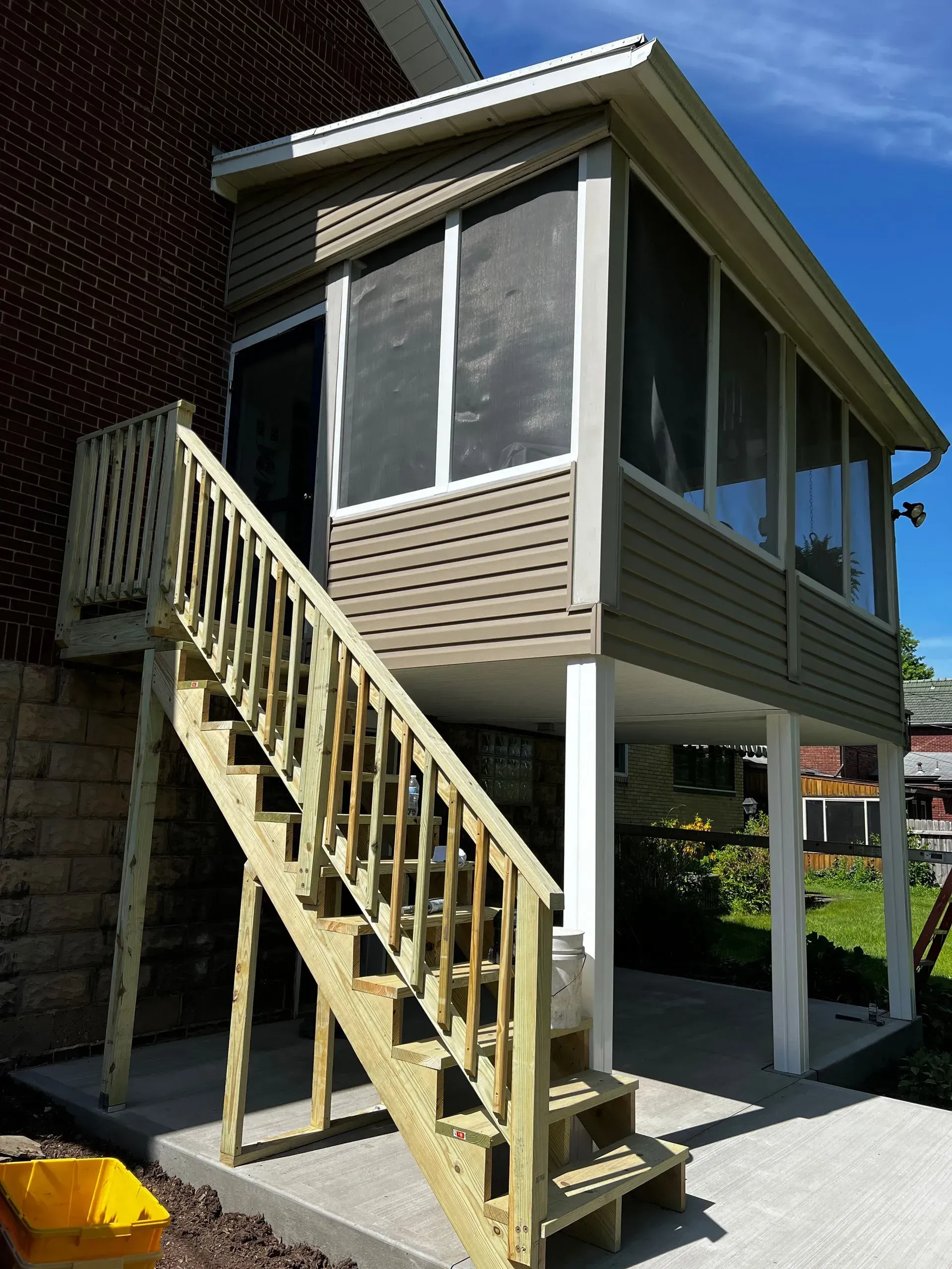 Screened-in porch with wooden stairs attached to a brick building. Porch has beige siding, white trim, and a concrete patio.