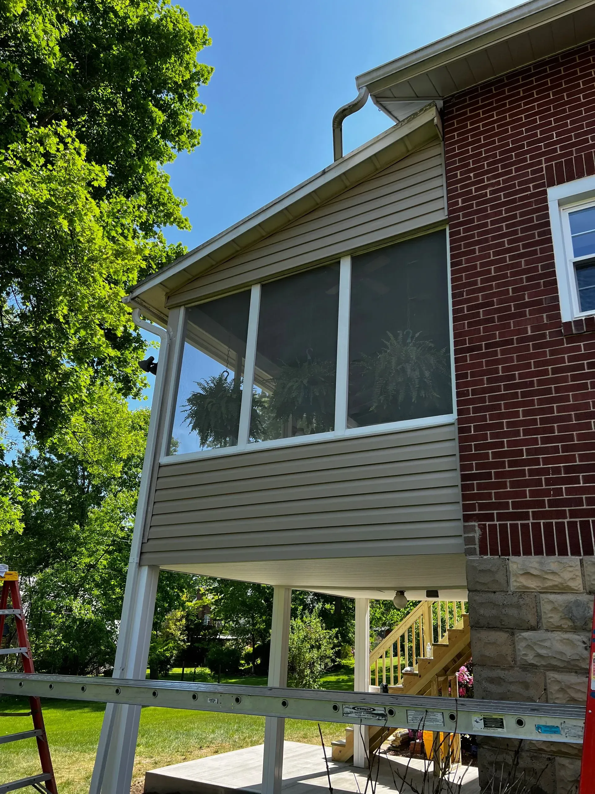 Screened porch attached to a brick house with tan siding, blue sky, and a ladder.