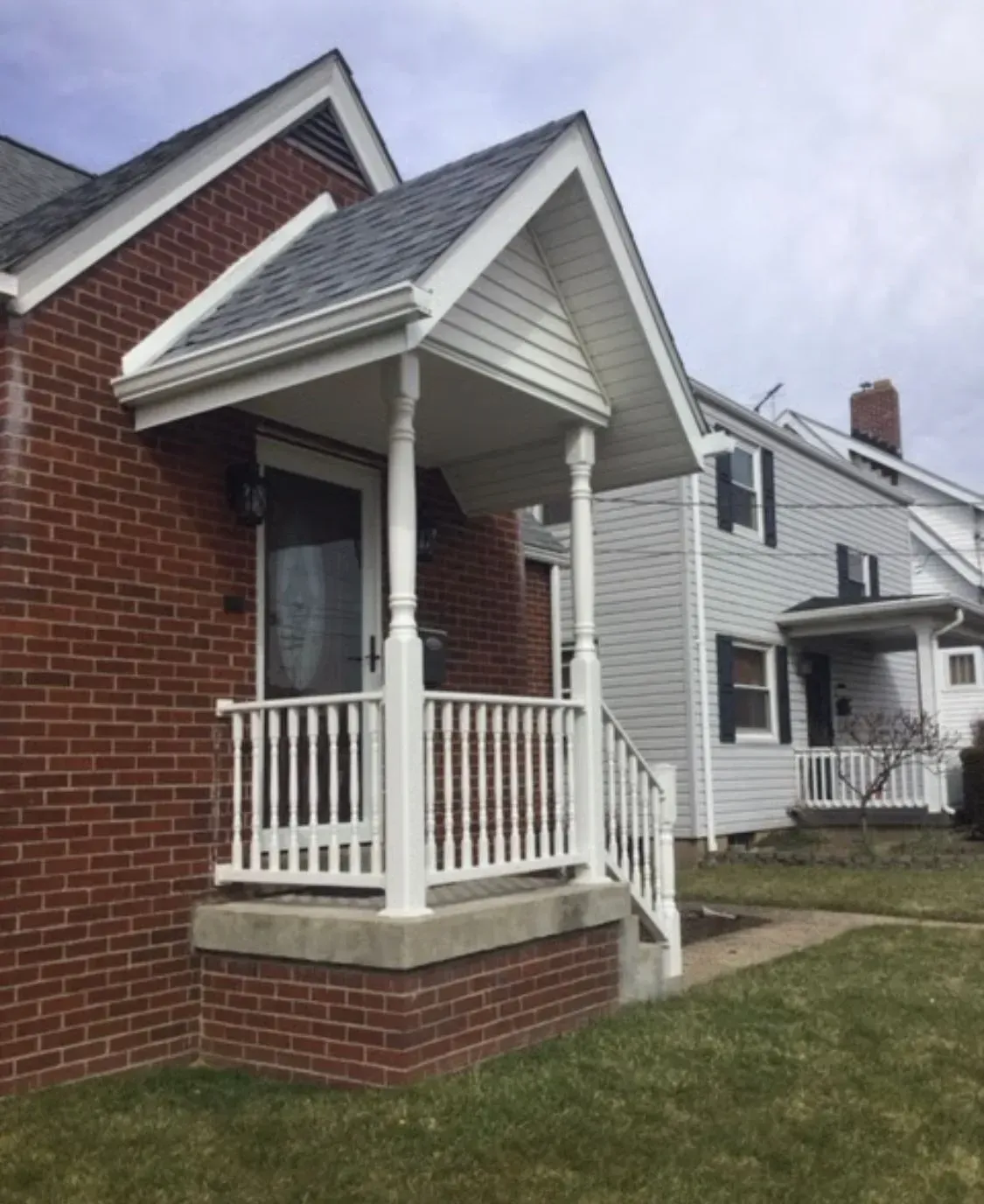 Brick house with a small porch, white railing, and a gray roof. Another house in the background.