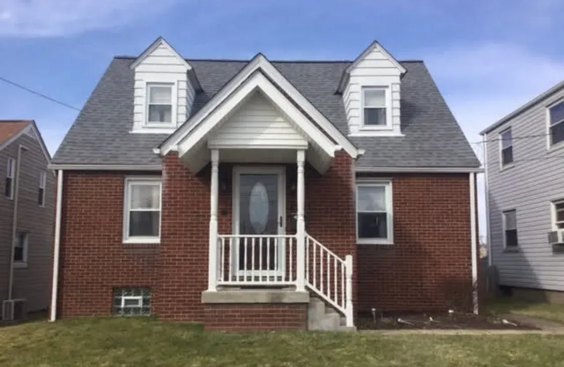 Red brick house with grey roof, two dormers, white trim, and porch, under a blue sky.