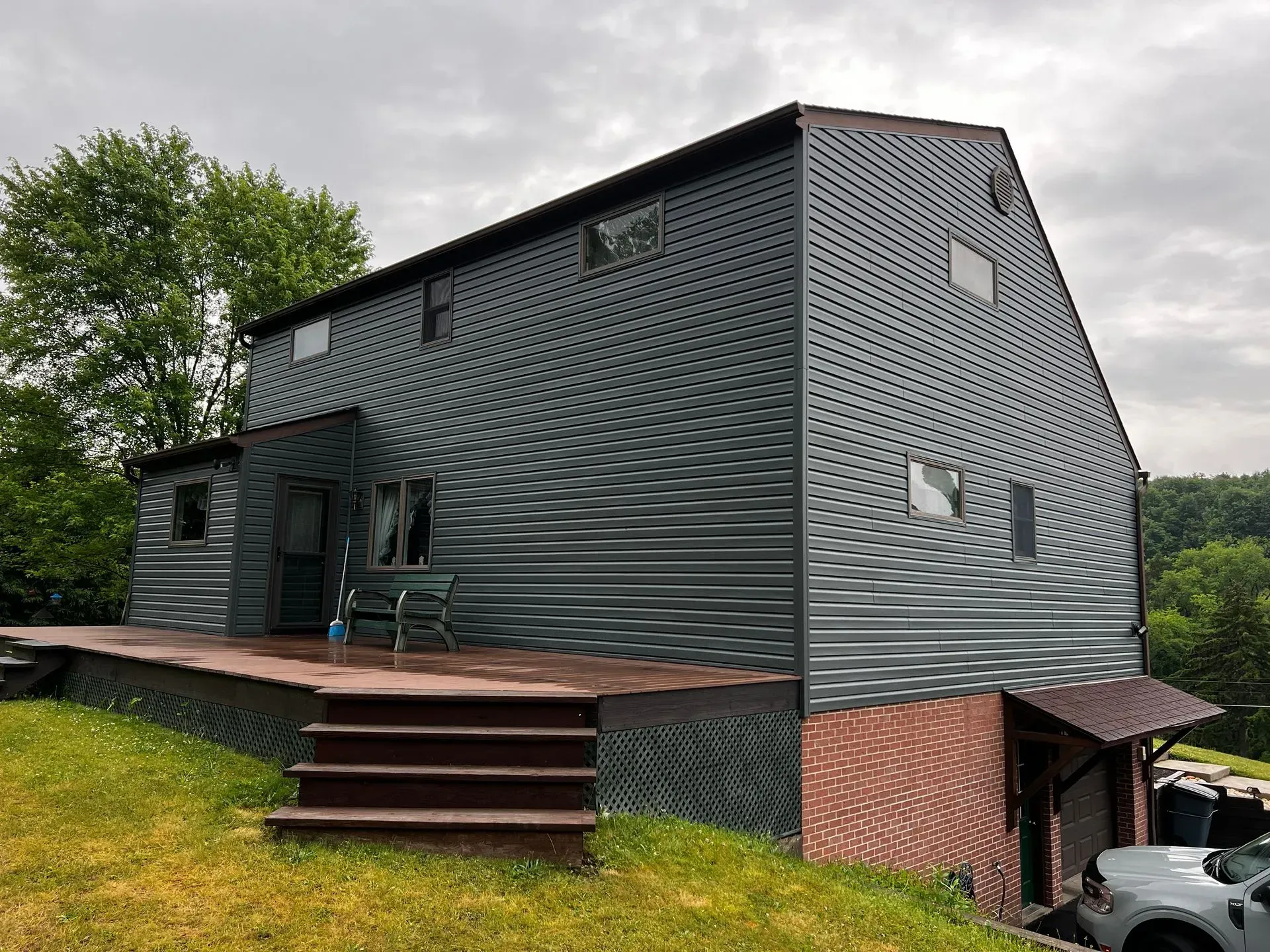 Two-story house with gray corrugated metal siding, a deck, and brick foundation. Overcast sky.