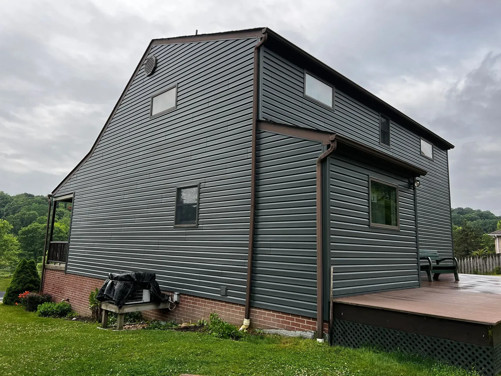 Dark gray house with brown trim and siding, a small deck, and a grassy yard under a cloudy sky.