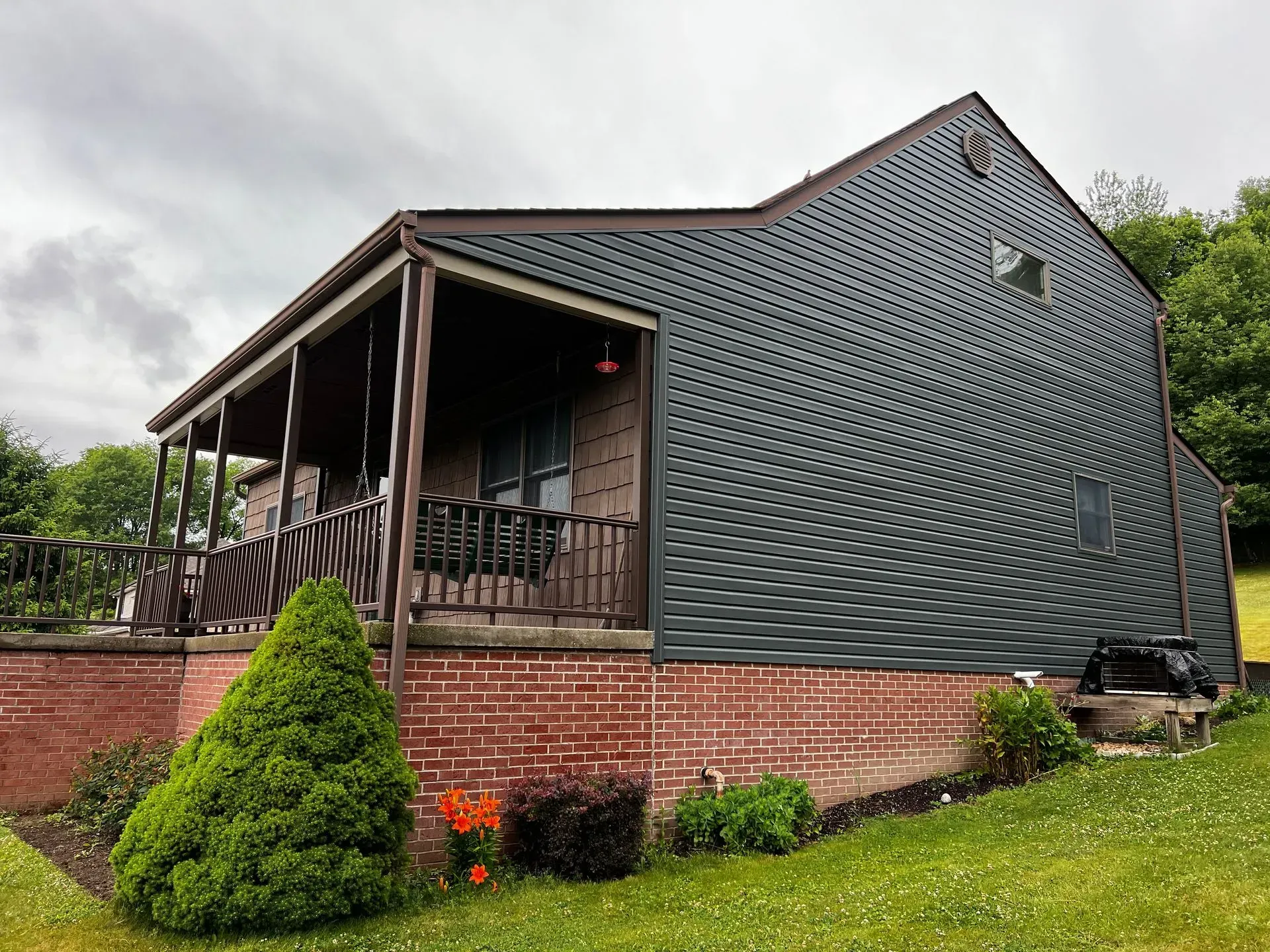 House with covered porch, brick base, dark gray siding, and green lawn under a cloudy sky.
