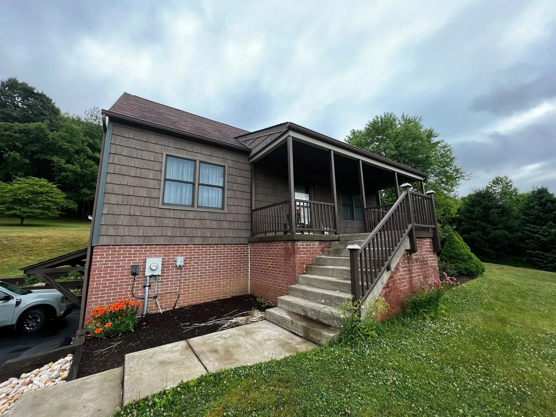 Brown house with a porch and steps, set in a grassy yard under a cloudy sky.
