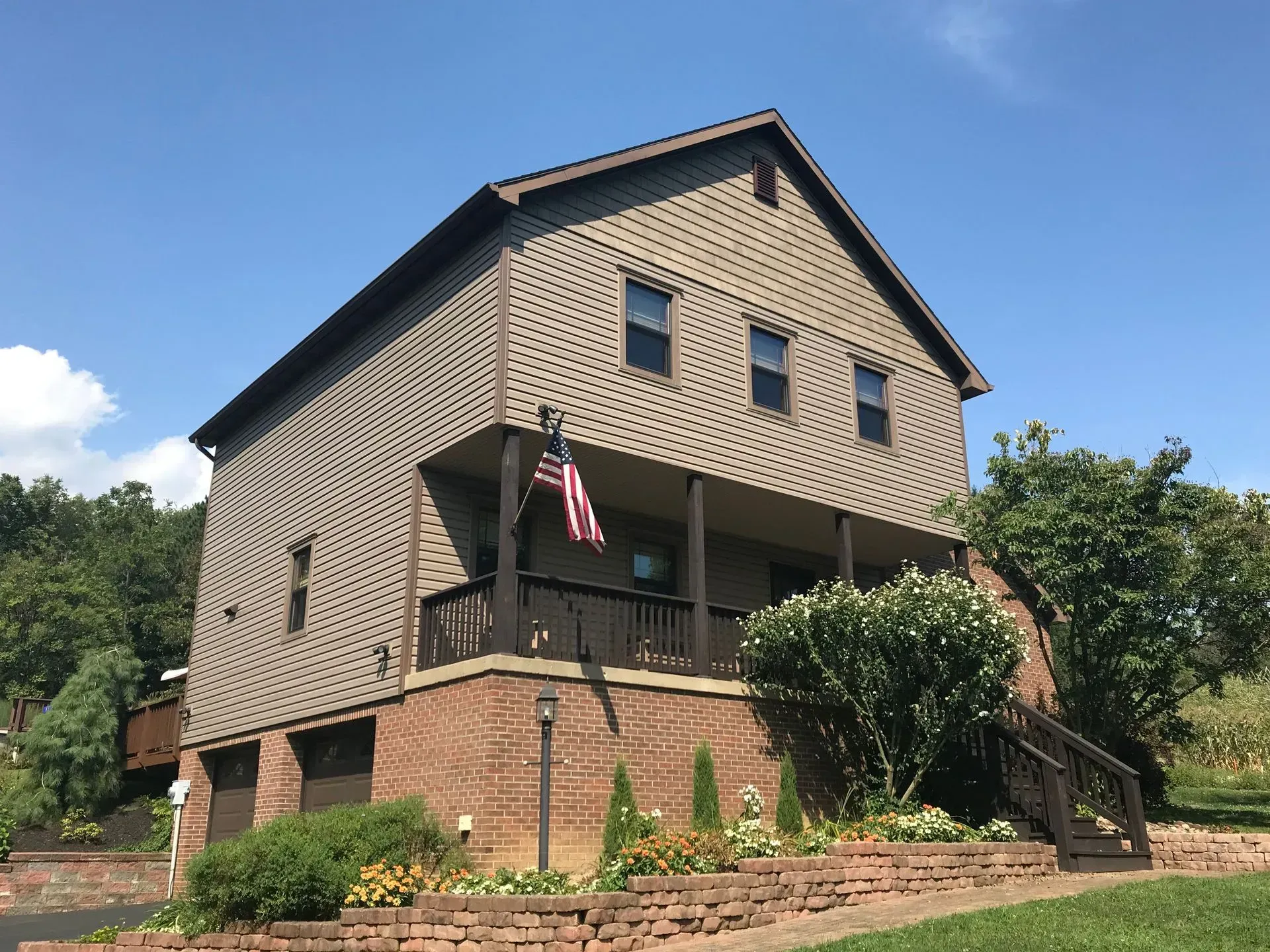 Two-story house with brown siding, porch, and American flag; brick base, landscaped front yard under a blue sky.
