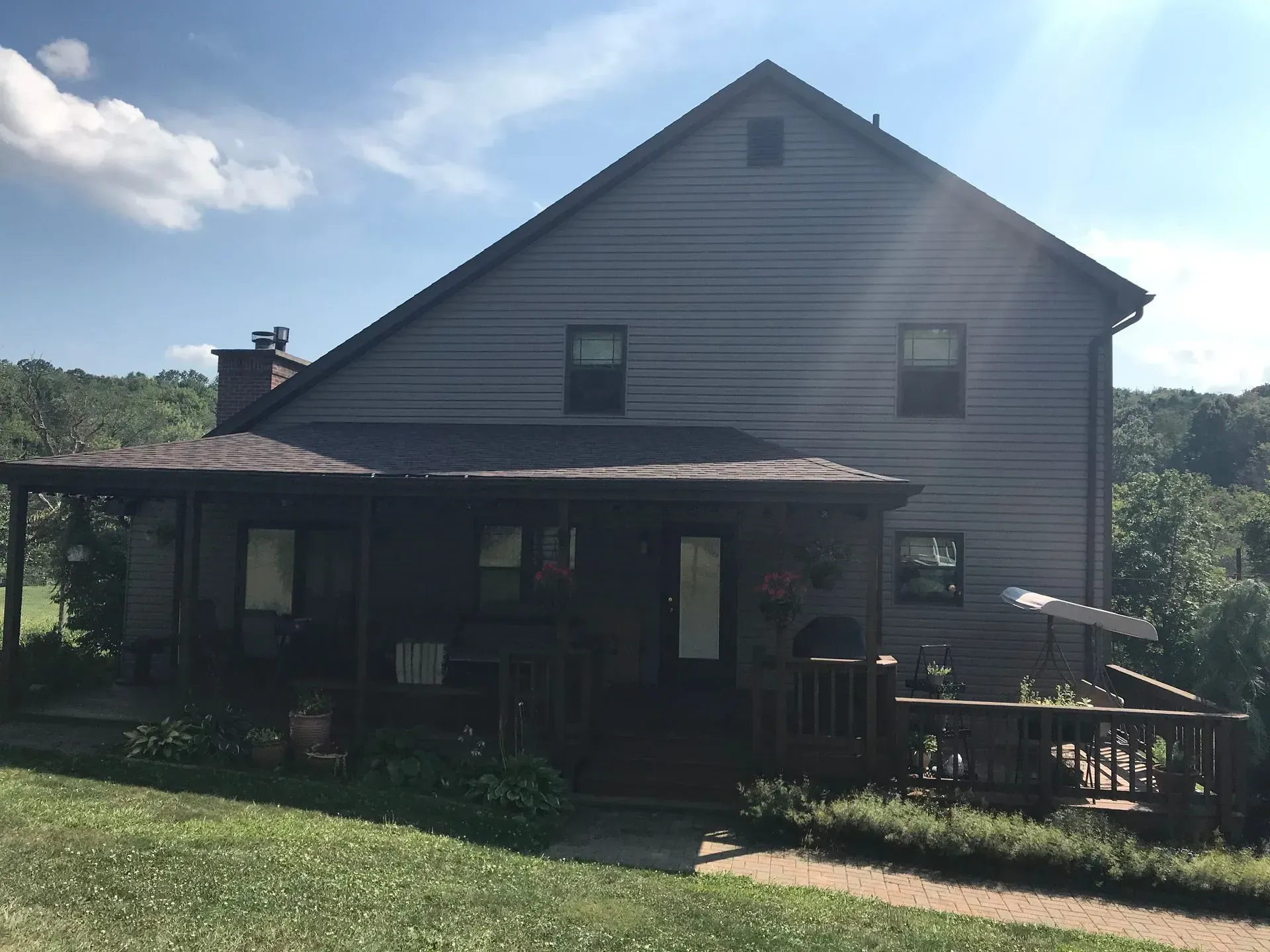 Two-story house with dark gray siding and a wraparound porch, set in a green landscape under a sunny sky.