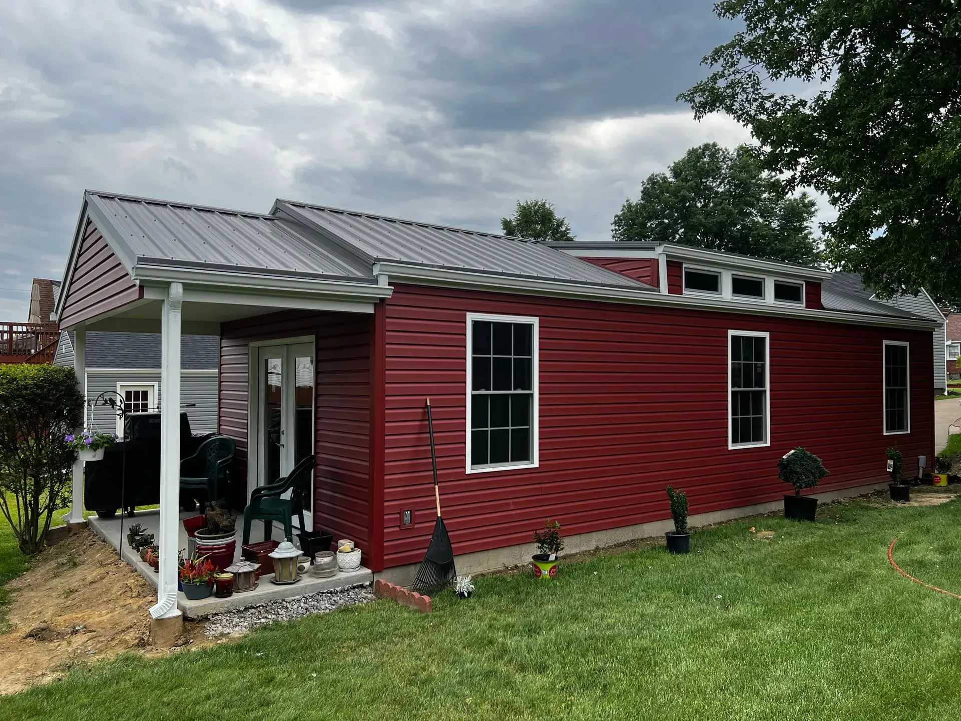 Red-sided building with a metal roof and a covered porch on a grassy hill under a cloudy sky.