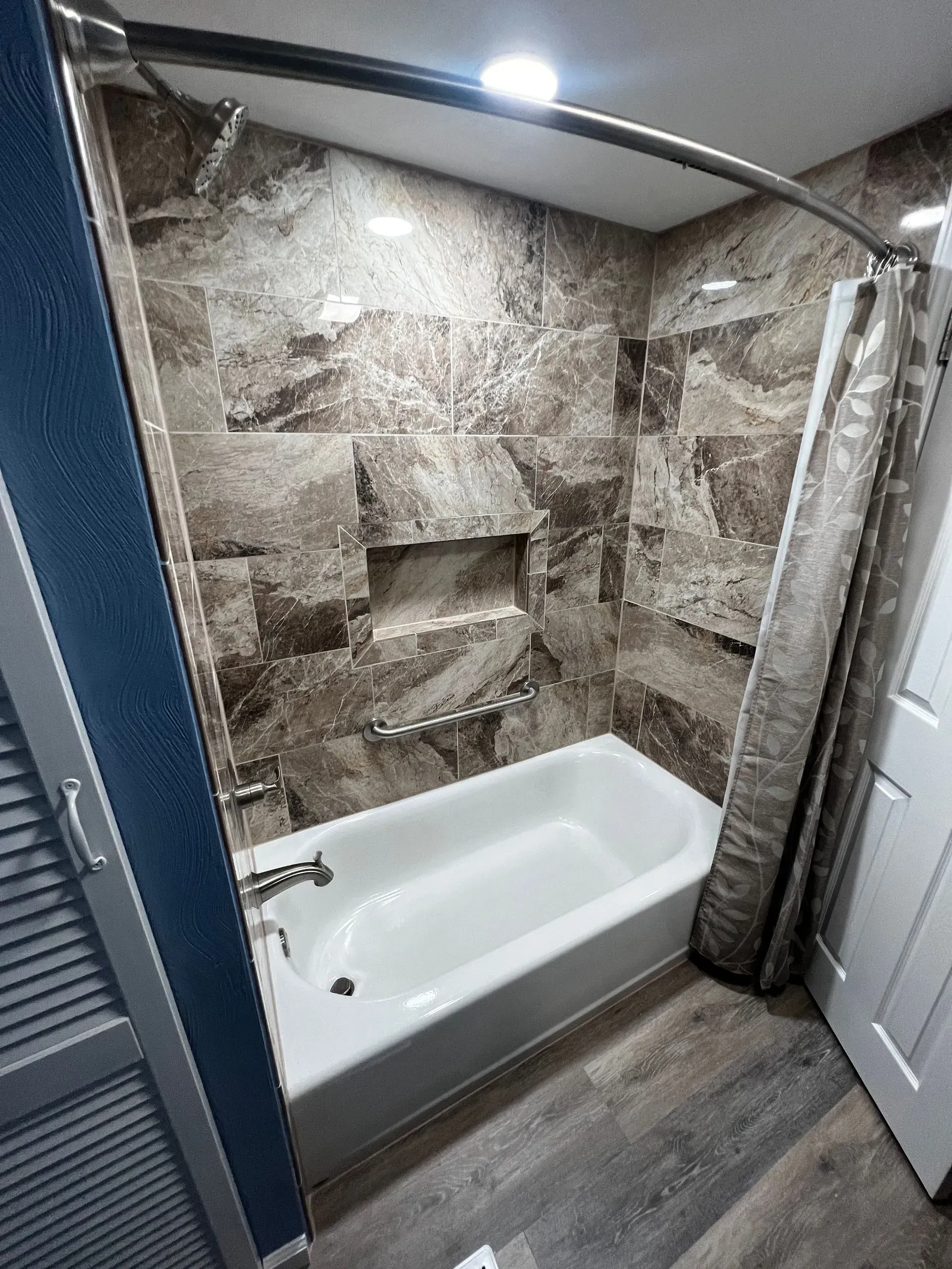 Bathroom with beige marbled tile, white bathtub, and wooden-look flooring.