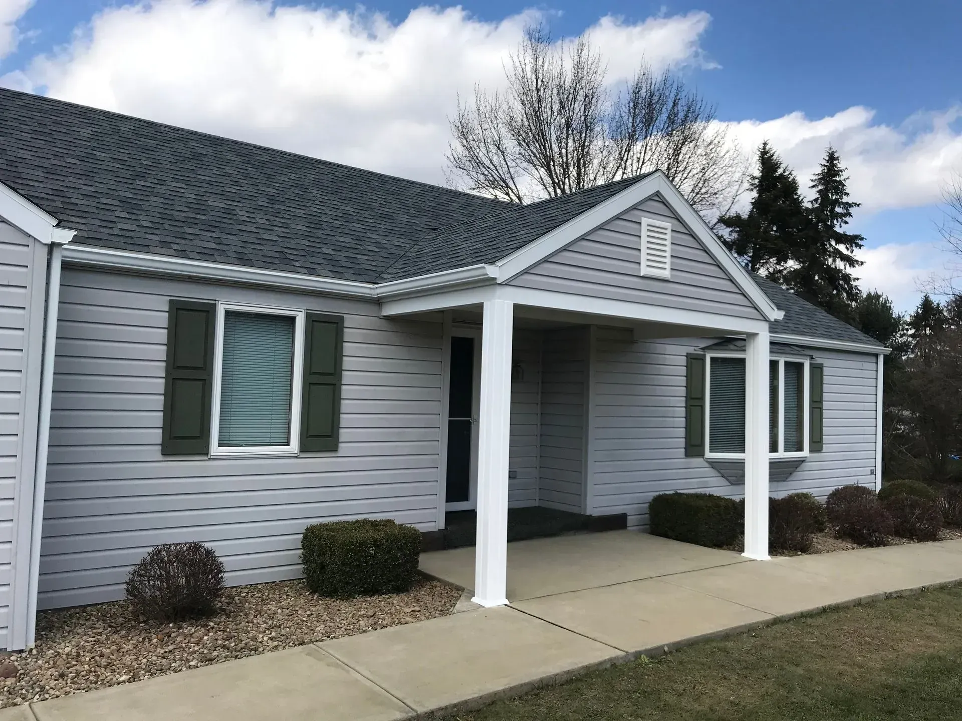 Light gray house with a dark gray roof and green shutters under a blue sky.