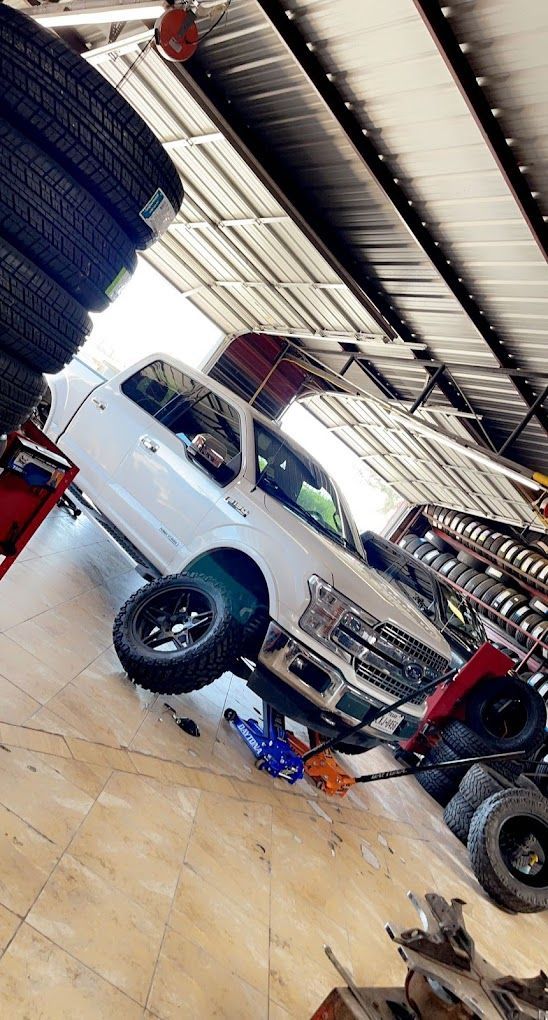 White truck on a jack in a garage, one wheel raised, surrounded by tires.