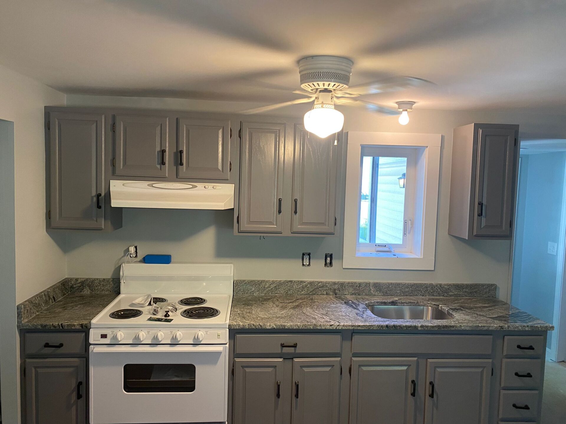 A kitchen with gray cabinets , a white stove , a sink and a ceiling fan.