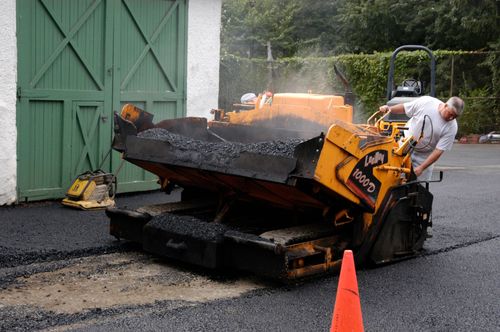 Worker using a yellow asphalt paver near a green garage door, with steam rising from fresh pavement.