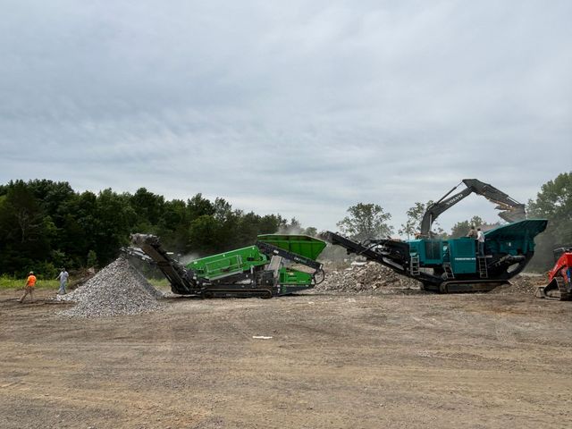 Two green and black demolition machines working on a gravel pile in a dirt lot under cloudy skies