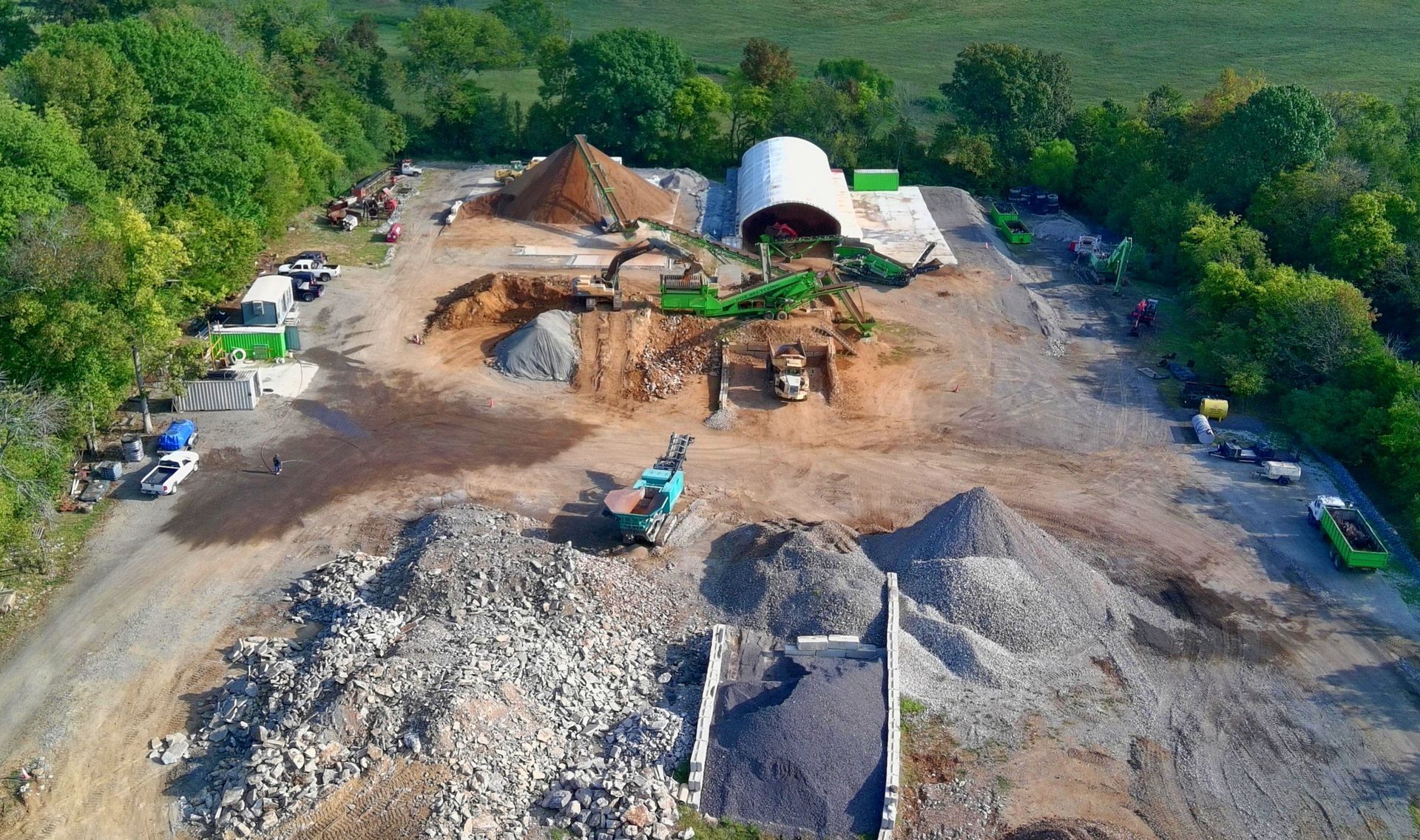 Aerial view of a construction site with dirt piles, machinery, and a large white covered structure surrounded by trees