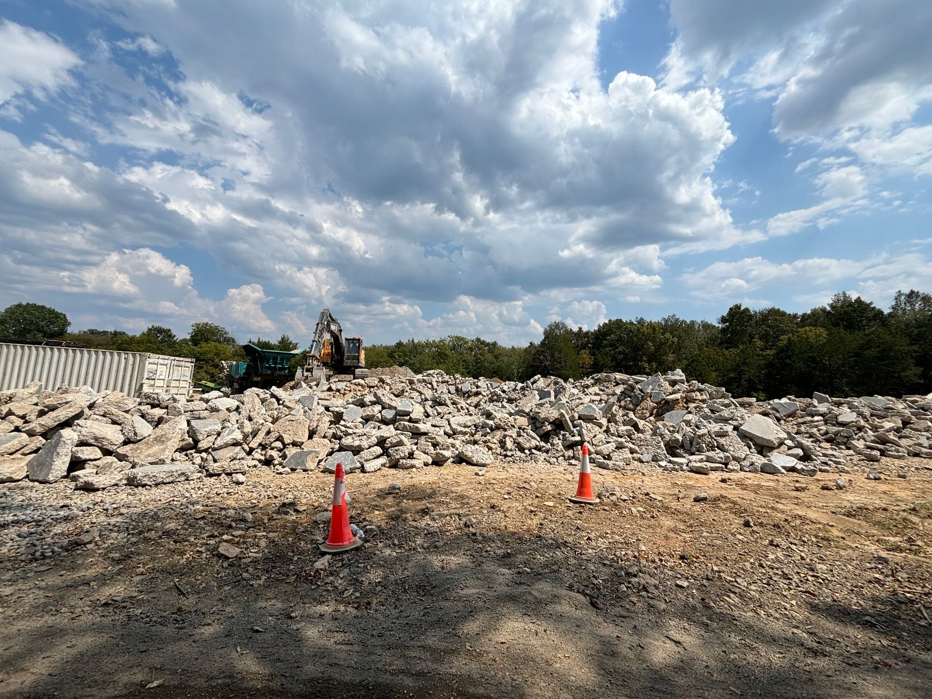 Construction site with rubble pile, excavator, and two orange traffic cones under a cloudy sky