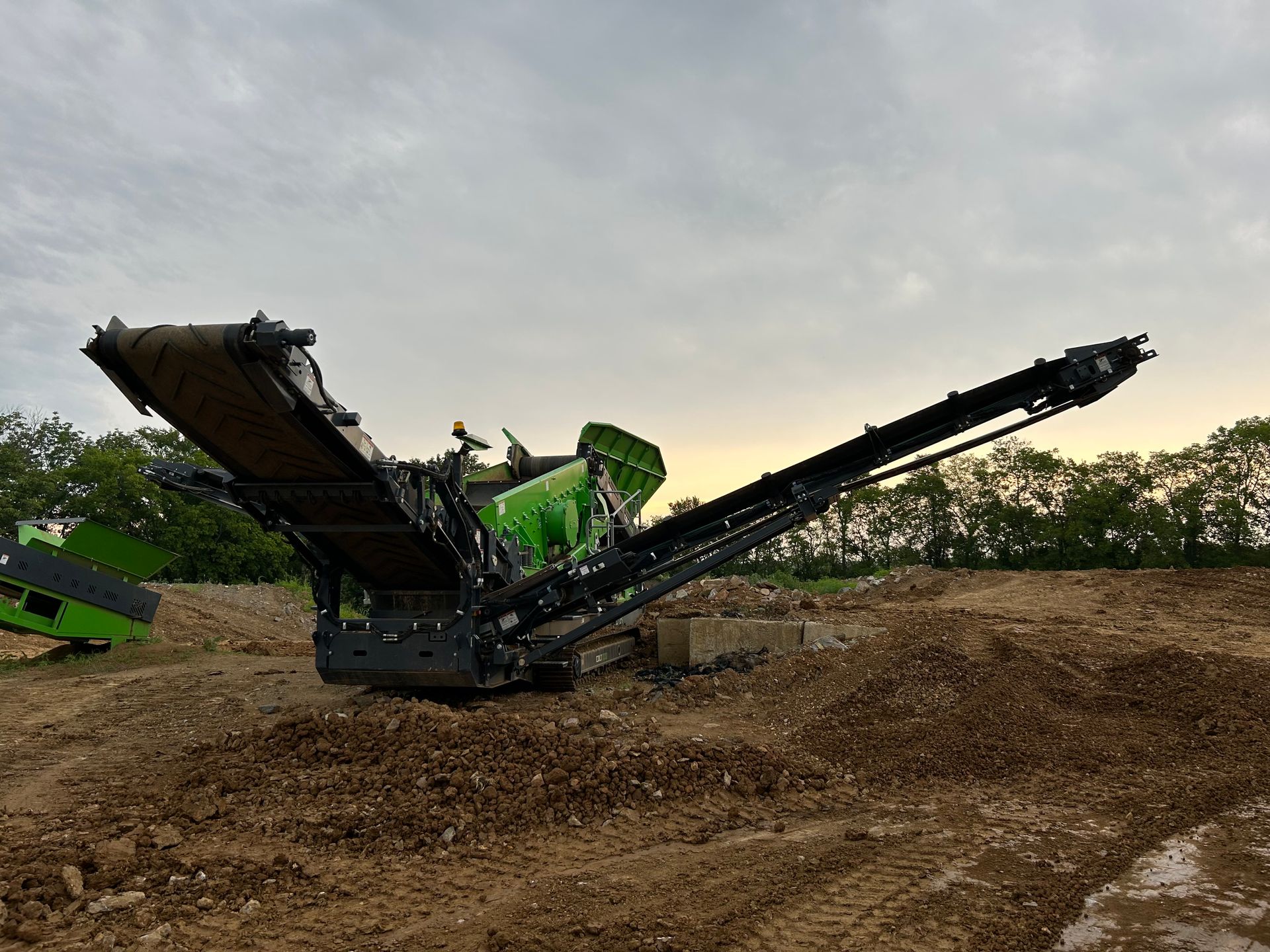 Green heavy-duty excavator with extended boom at a dirt quarry under a cloudy sky
