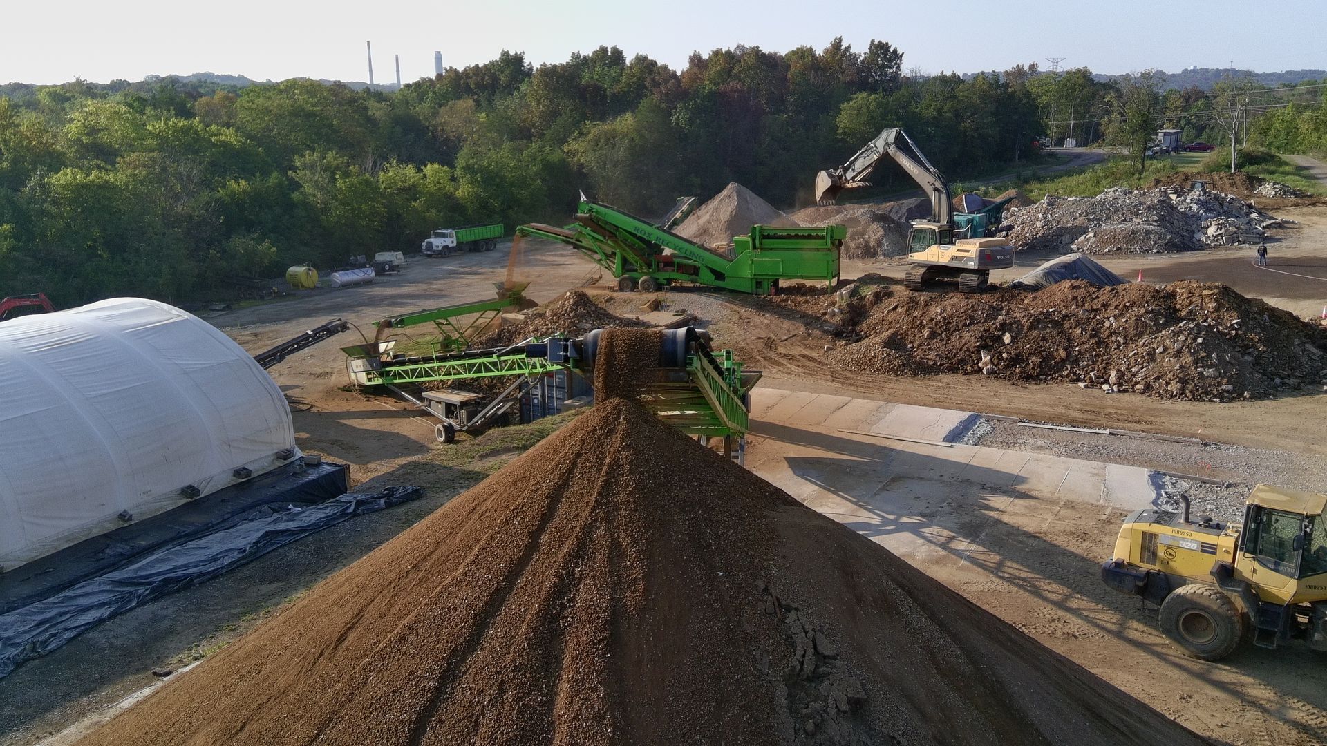 Aerial view of a construction site with gravel piles, excavators, trucks, and a white storage tent.