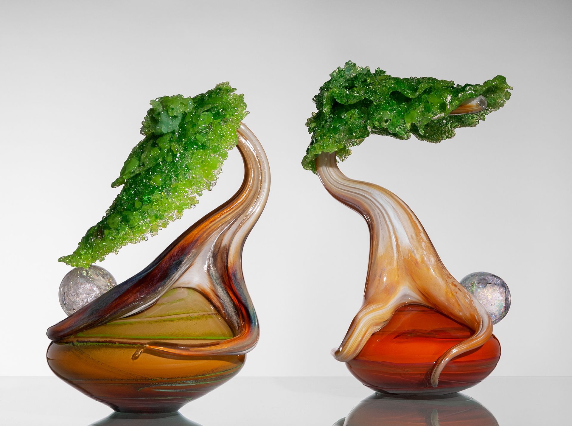 Two abstract glass vases with green leafy tops, one amber and one orange, on a white background