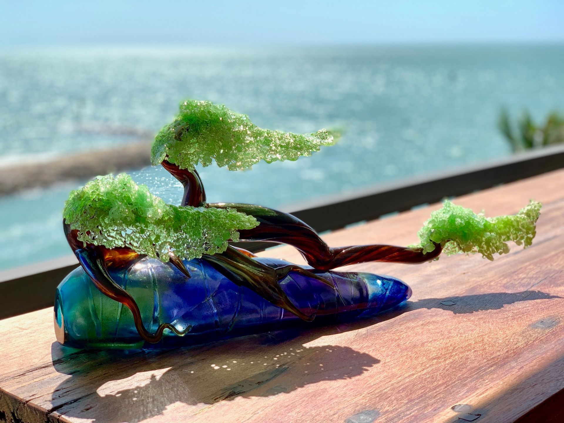 Small bonsai tree growing from a blue glass shoe on a wooden railing by the sea