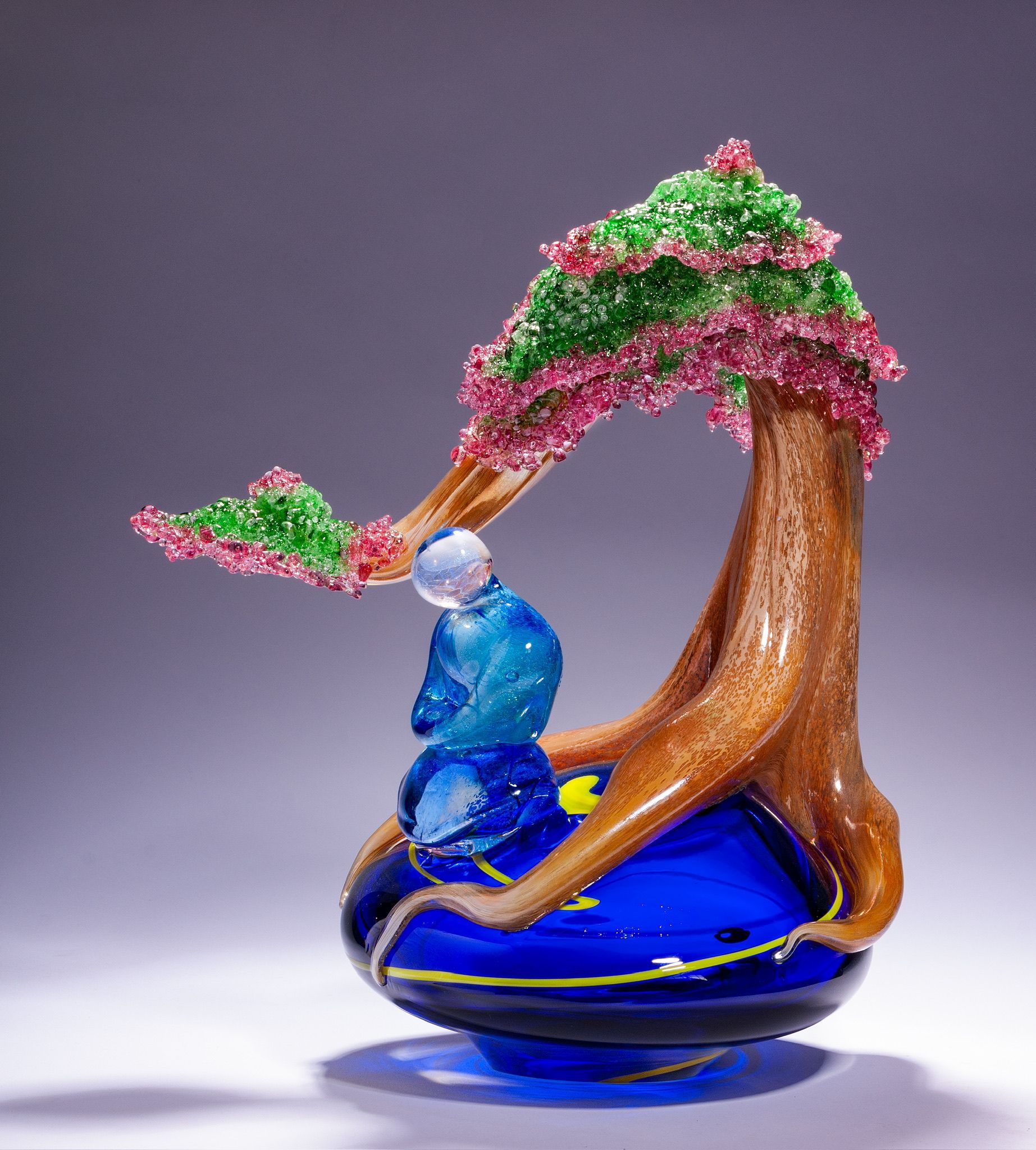 Glass art sculpture of a blue figure beneath a brown tree with pink and green foliage on a blue base