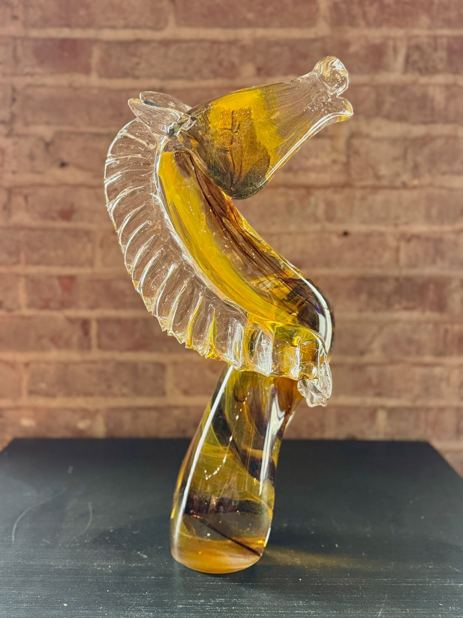 Amber glass sculpture with a curved, ribbed form on a black surface against a brick wall