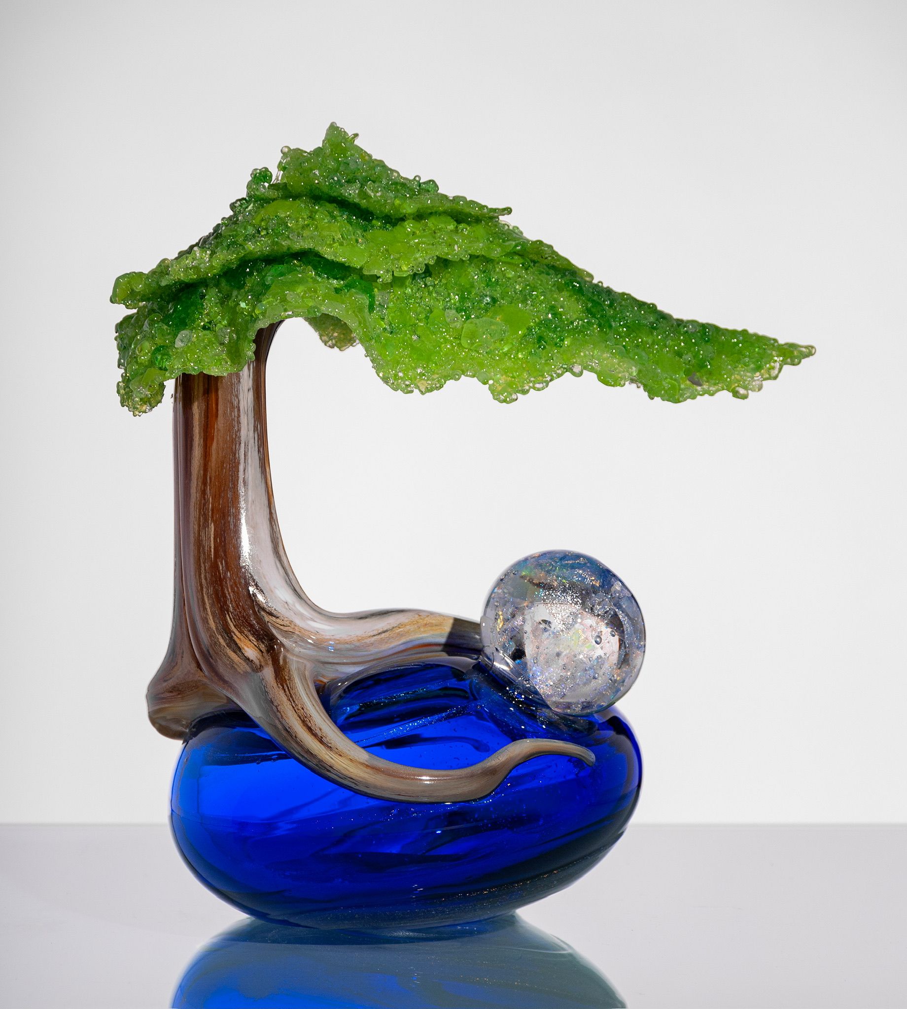 Glass tree sculpture with green canopy, brown trunk, and blue base on a white background