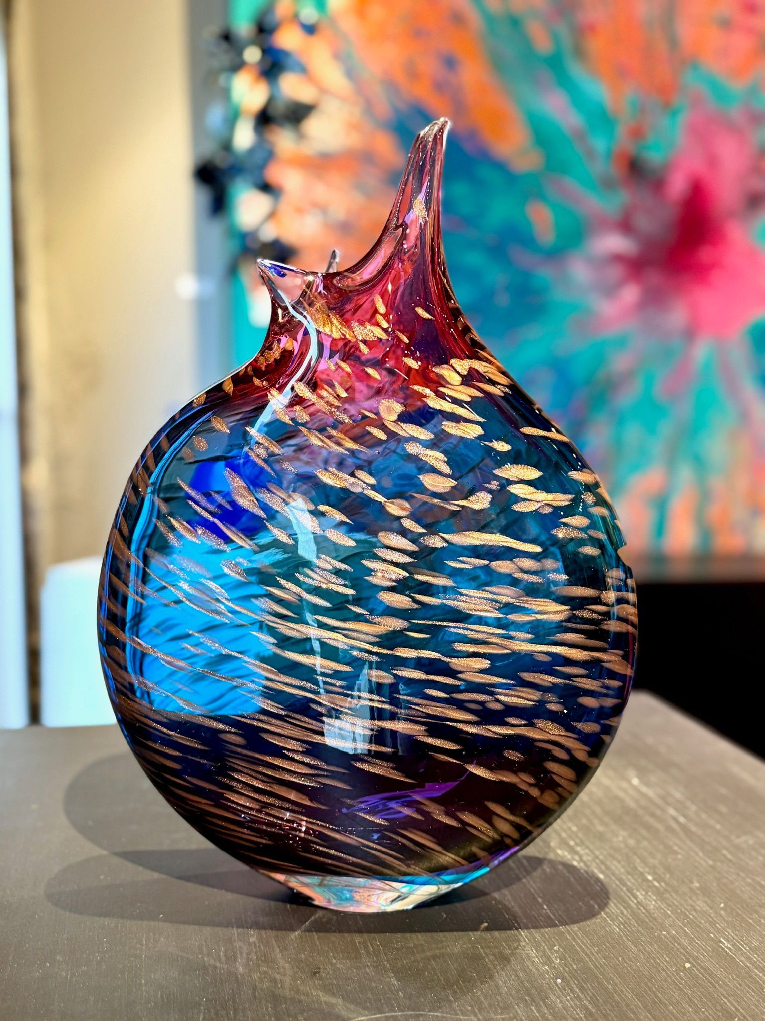 Colorful glass sculpture with a pointed top, blue and gold swirls on a tabletop, blurred art in the background