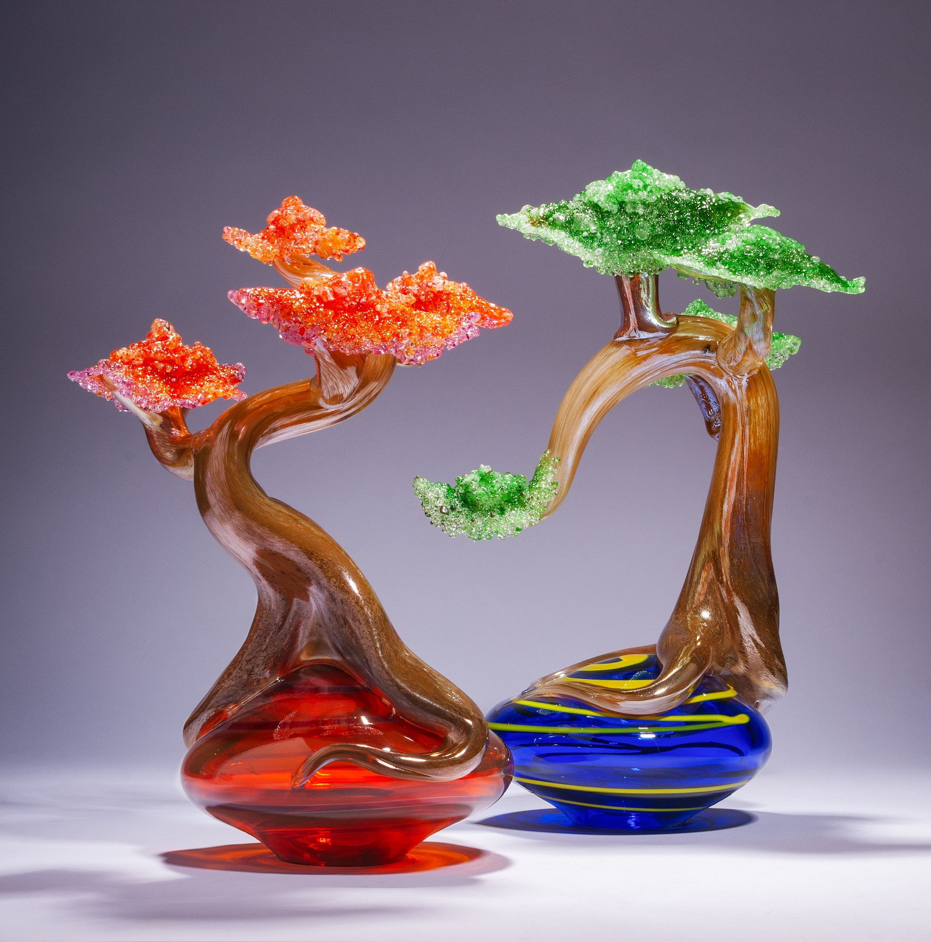 Two small glass bonsai trees, one red-orange and one green-blue, on a neutral background.