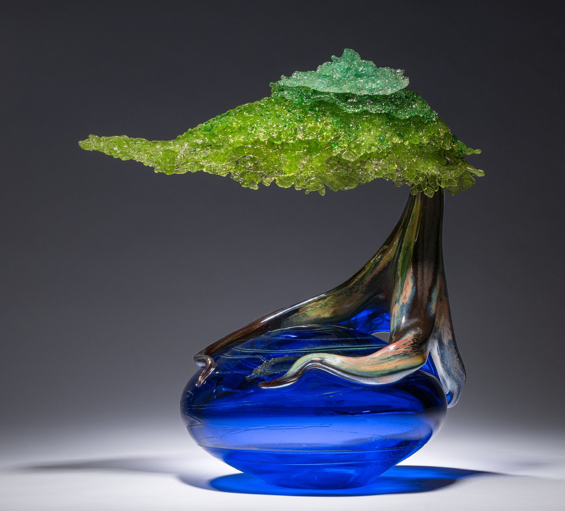 Glass sculpture of a blue droplet-shaped vase with a green leaf-like top on a gray background