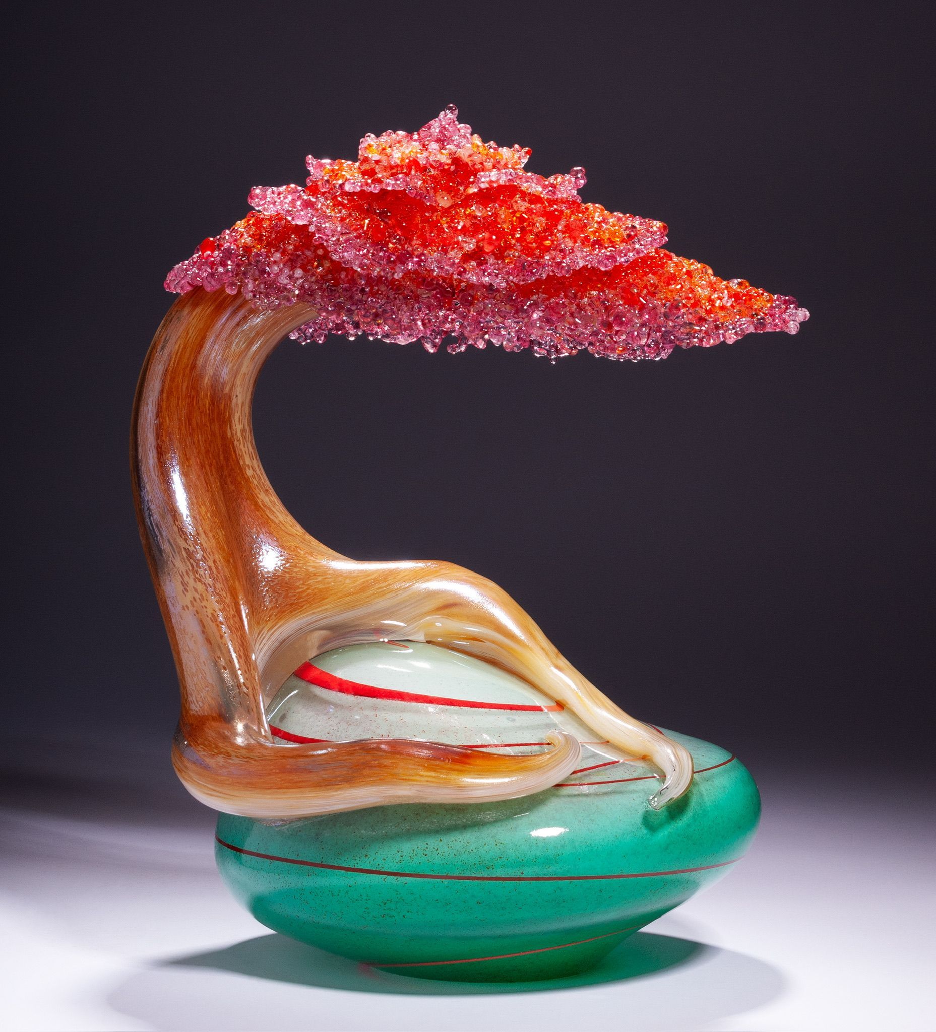 Glass sculpture of a reclining figure beneath a red tree canopy on a green base