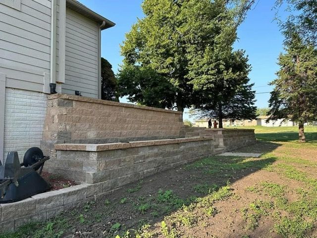 Brick retaining wall beside a house with a grassy yard and large trees in the background