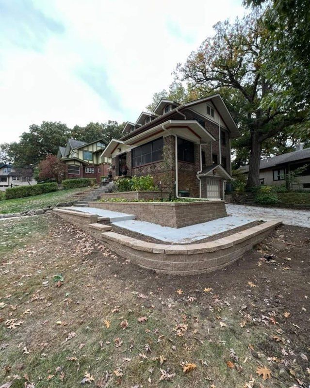 Stone house with a sloped roof and covered porch in a wooded yard, viewed from the front lawn.