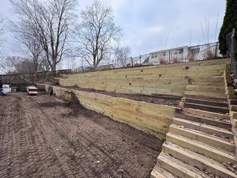 Terraced dirt slope with wooden steps and retaining walls beside a muddy lot and leafless trees