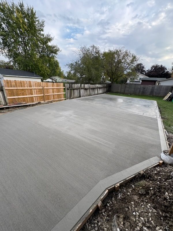 Freshly poured concrete slab in a fenced backyard with trees and cloudy sky