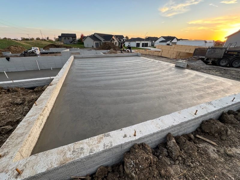 Fresh concrete slab foundation at a construction site during sunset, with houses and trucks in the background