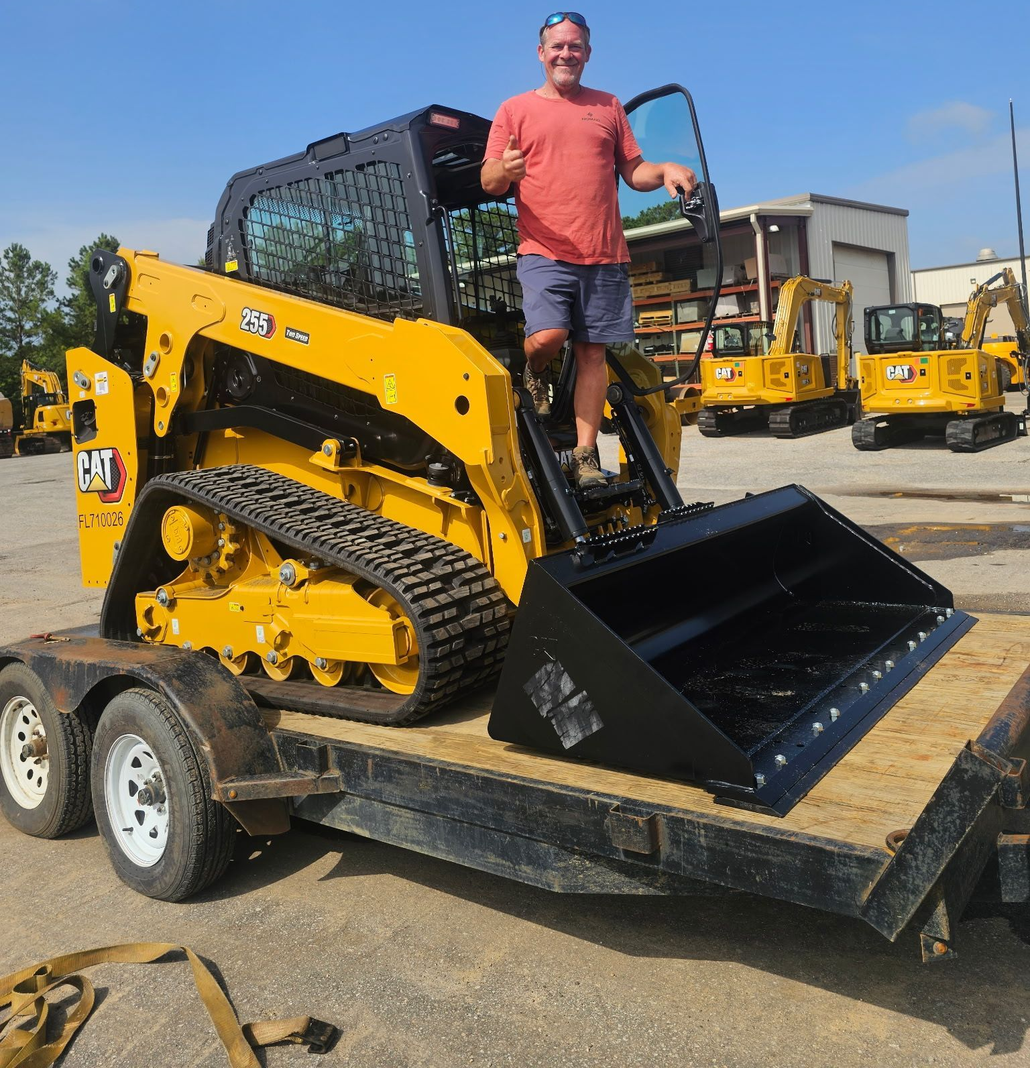Man standing on a yellow Caterpillar skid steer on a trailer, thumbs up, sunny outdoor setting.