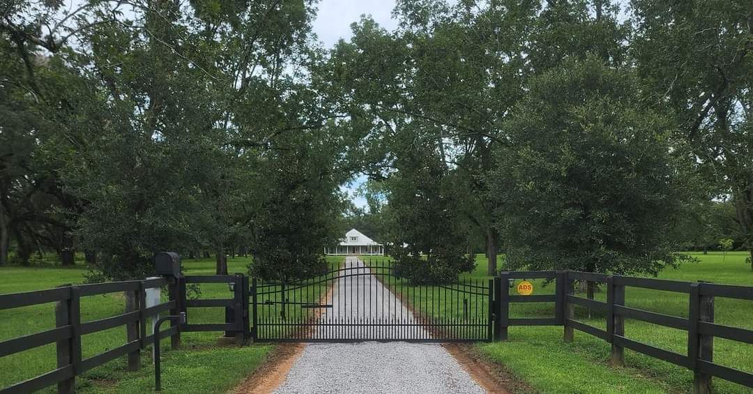Gravel driveway with closed gate leads to a house. Trees border the drive, with a wooden fence on each side.