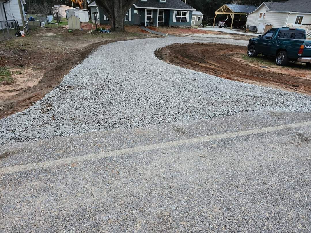 Gravel driveway curving toward a house, dirt border on sides; a pickup truck is parked nearby.