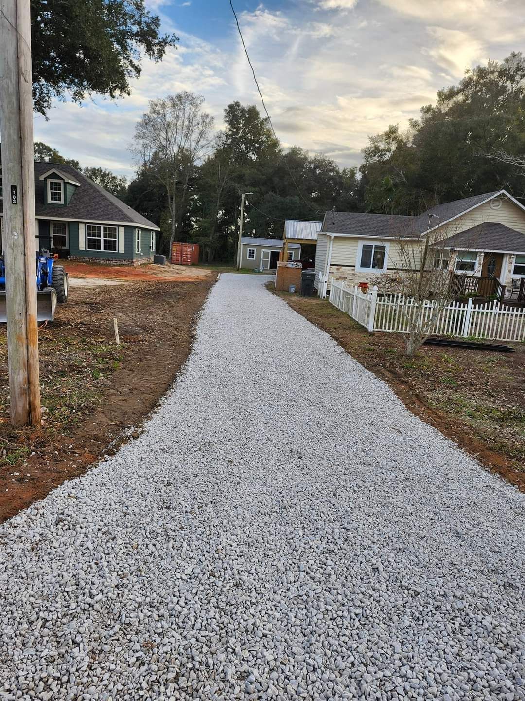 Gravel driveway leads to houses on a sunny day; a utility pole is on the left.