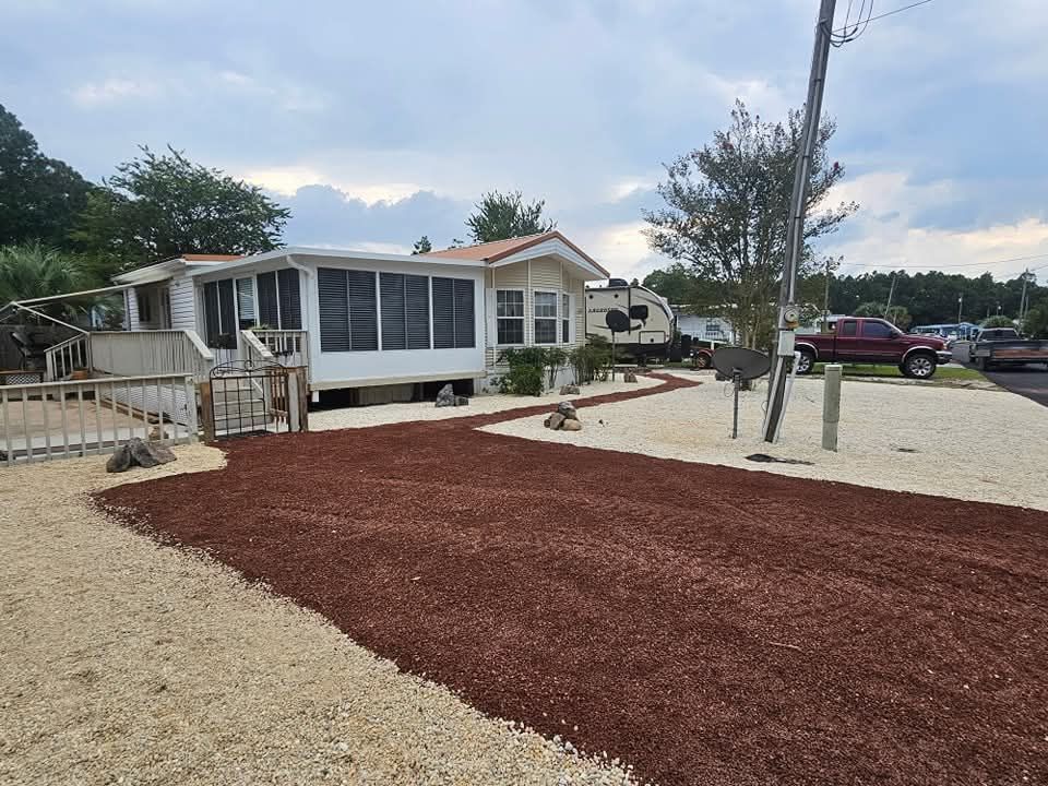 White mobile home with porch, walkway, and red/beige gravel driveway, parked camper and red pickup truck.
