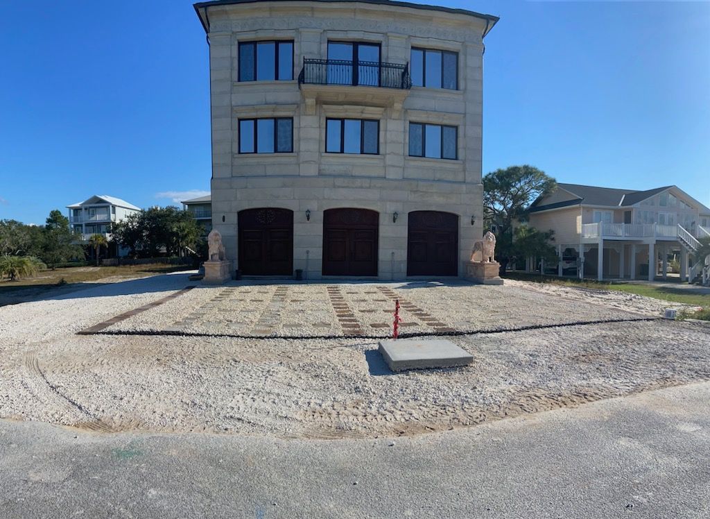 Three-story tan house with three garage doors. White gravel driveway, blue sky.