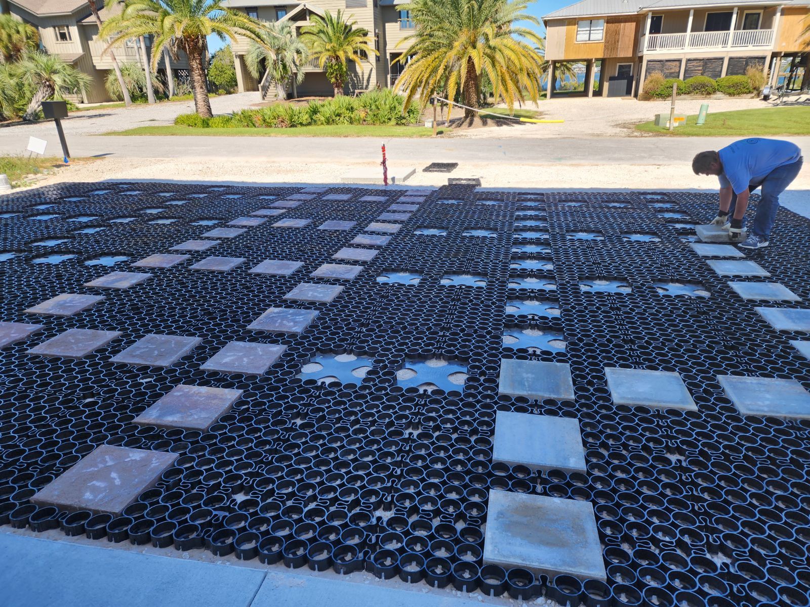 A man installing pavers in a driveway with a black grid, beach houses in the background.
