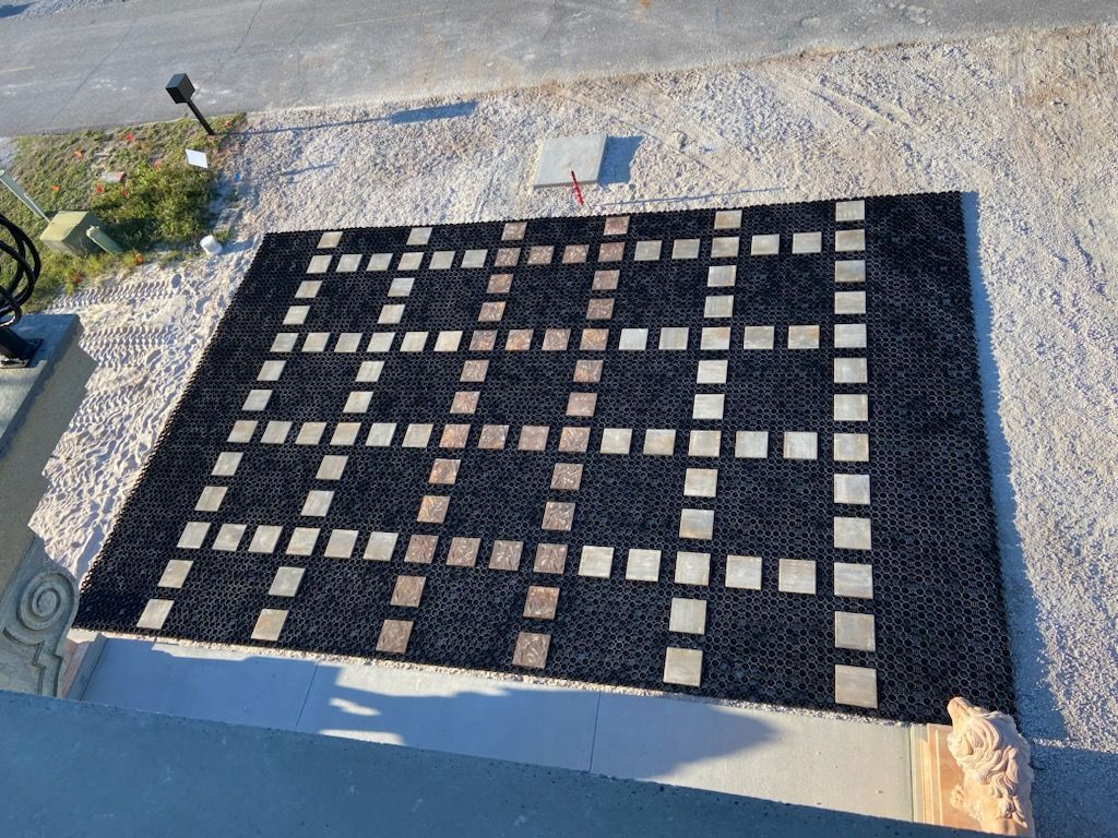 Black rug with a grid pattern of beige squares on a gravel surface.